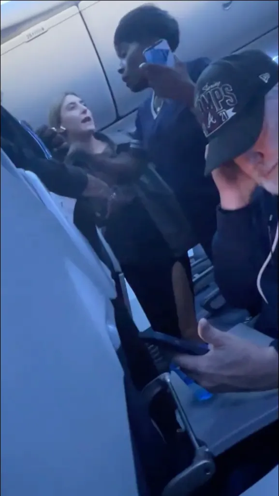 A flight attendant holding a phone to her ear and another passenger arguing in an airplane cabin.